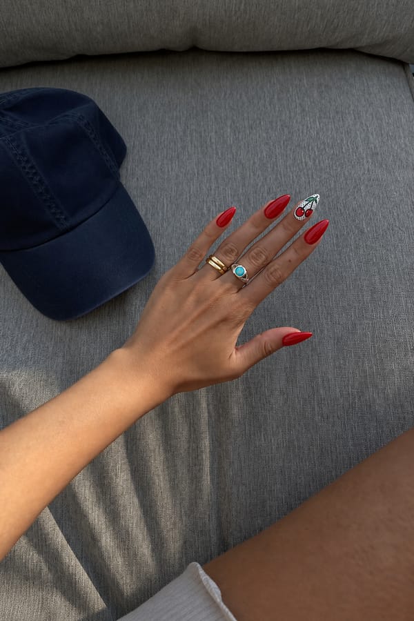 Bold cherry red glossy finish on medium almond nails, with one statement nail hand-painted with a retro comic-style pop art cherry motif in black outline. Nail shape: almond. Nail length: medium. Only change the nails visible in the image to match this design. Keep the rest of the image — the hand, skin, background, lighting, and everything else — exactly the same.