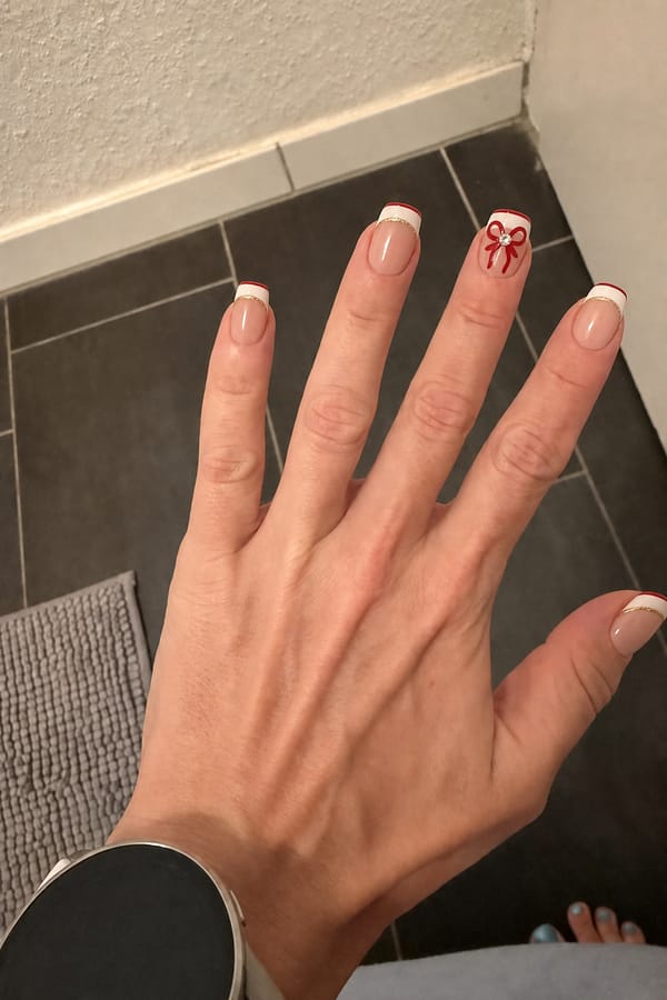 Glossy sheer nude base nails. Index, middle, and pinky fingers feature deep, thick light cream French tips. At the very top edge of these tips is a thin cherry red line. those fingers with a thin crisp gold outline underneath the French tip framing the smile line. The ring finger has no French tip and no gold line; it only features the thin cherry red line along the edge and a big hand painted cherry red bow with a rhinestone in the center of the bow. Nude base is clean. Modern, ultra-precise. Nail shape: square. Nail length: medium. Only change the nails visible in the image to match this design. Keep the rest of the image — the hand, skin, background, lighting, and everything else — exactly the same.