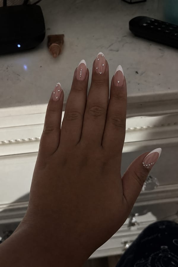 Glossy sheer nude base with crisp white French tips. All 5 nails are accent designs. Each nail features small silver rhinestones placed along the center or near the tip. Variation includes: some nails with classic French tips + scattered rhinestones, some with extended white tips + single rhinestone near the edge, and some nude areas with minimal spaced rhinestones only. Nail shape: almond. Nail length: medium. Only change the nails visible in the image to match this design. Keep the rest of the image — the hand, skin, background, lighting, and everything else — exactly the same.