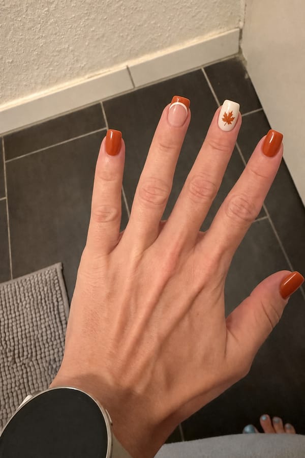 Glossy burnt orange nails. ring finger matte creme white nails with ring finger with a hand painted maple leaf in burnt orange with chestnut brown details. middle finger glossy bide base with burnt orange French tip thin crisp creme white outline underneath framing the smile line the line. Nail shape: square. Nail length: medium. Only change the nails visible in the image to match this design. Keep the rest of the image — the hand, skin, background, lighting, and everything else — exactly the same.