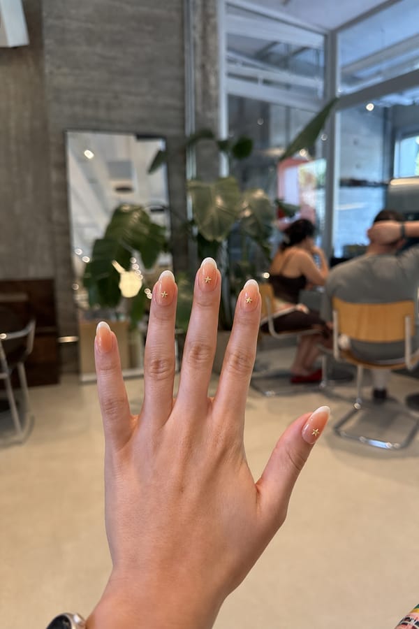Sheer nude glossy base with a soft orange-to-white radial gradient (aura effect) concentrated toward the tips. A mix of styles across the set: some nails feature a smooth gradient only, while others include blended white French tips layered over the gradient. Accent details on 4 nails: 3 nails have small gold star decals placed slightly above the tip area, centered or slightly offset. 1 nail has a gold star decal placed lower toward the mid-area of the nail. Nail shape: almond. Nail length: medium. Only change the nails visible in the image to match this design. Keep the rest of the image — the hand, skin, background, lighting, and everything else — exactly the same.