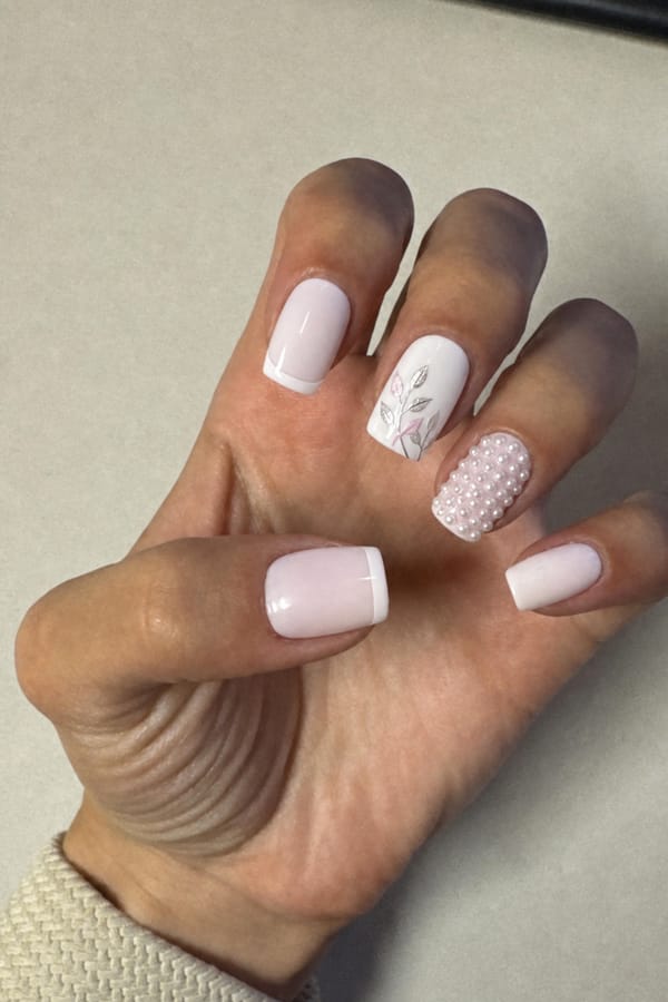 Milky white base on two nails and white crisp French tip. two nails full white with a leaves in silver and baby pink. another nail with the same baby pink and lots of white tiny pearls all over. Nail shape: square. Nail length: medium. Only change the nails visible in the image to match this design. Keep the rest of the image — the hand, skin, background, lighting, and everything else — exactly the same.