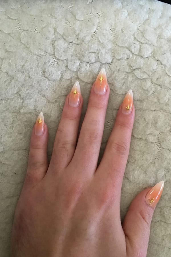 Sheer nude glossy base with soft orange-to-white radial gradient (aura) focused toward the tips. Some nails have blended white French tips over the gradient. 4 accent nails: 3 with small gold star decals near the tip, 1 with a gold star placed slightly lower. Remaining nails are gradient-only or gradient + soft French blend. Nail shape: stiletto. Nail length: long. Only change the nails visible in the image to match this design. Keep the rest of the image — the hand, skin, background, lighting, and everything else — exactly the same.
