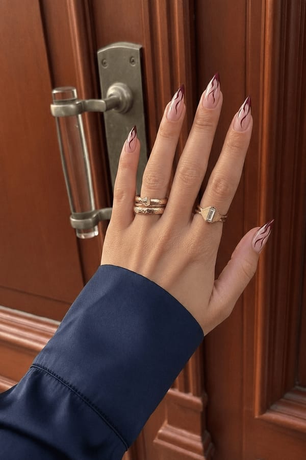 Sheer nude base with deep burgundy abstract flame-like tips and thin flowing line details. Bold, edgy, minimal aesthetic, high-gloss finish. Nail shape: stiletto. Nail length: medium. Only change the nails visible in the image to match this design. Keep the rest of the image — the hand, skin, background, lighting, and everything else — exactly the same.