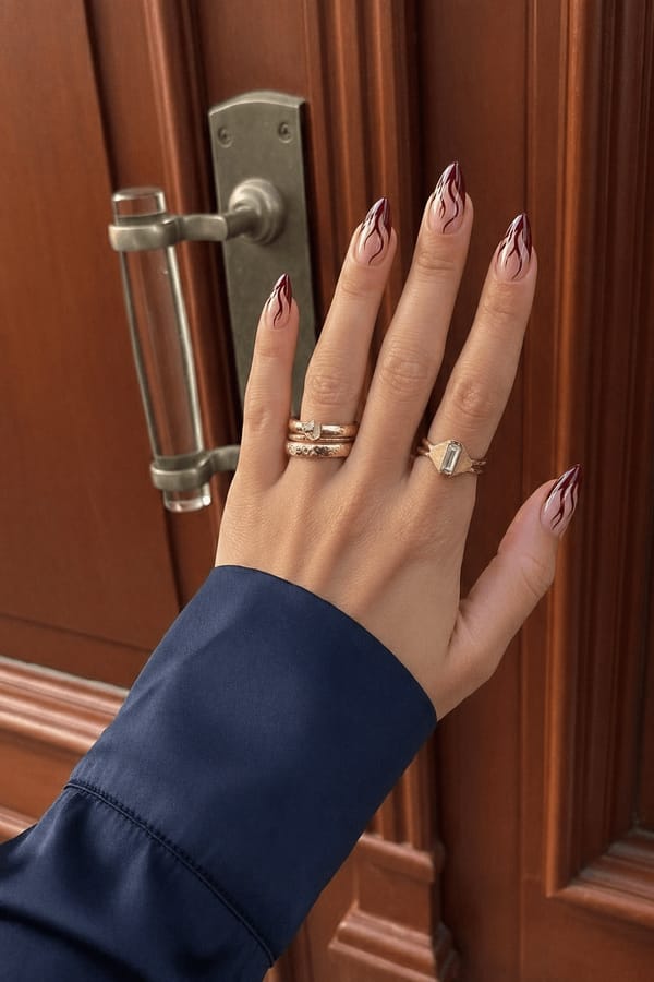 Sheer nude base with deep burgundy abstract flame-like tips and thin flowing line details. Bold, edgy, minimal aesthetic, high-gloss finish. Nail shape: stiletto. Nail length: medium. Only change the nails visible in the image to match this design. Keep the rest of the image — the hand, skin, background, lighting, and everything else — exactly the same.