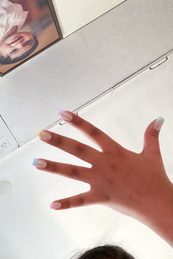 Glossy sheer nude base with pastel French tips. 5 accent nails: each nail features a different soft pastel tip color (lavender, yellow, peach, pink, mint, baby blue tones). Tips are clean and minimal with no outlines or decorations. All nails share the same style, only color varies per nail. Nail shape: square. Nail length: medium. Only change the nails visible in the image to match this design. Keep the rest of the image — the hand, skin, background, lighting, and everything else — exactly the same.