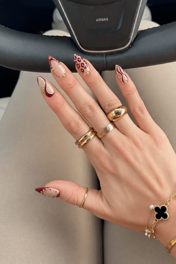 Milky nude base with burgundy accents and gold studs. All 5 nails are accent designs. One nail has burgundy French tip with gold studs. One has burgundy leopard print tips. Two nails feature burgundy abstract zebra/wave lines over nude base. One nail mixes soft burgundy patches with dots. Small gold bead clusters placed near cuticle or along patterns on each nail. Nail shape: stiletto. Nail length: medium. Only change the nails visible in the image to match this design. Keep the rest of the image — the hand, skin, background, lighting, and everything else — exactly the same.