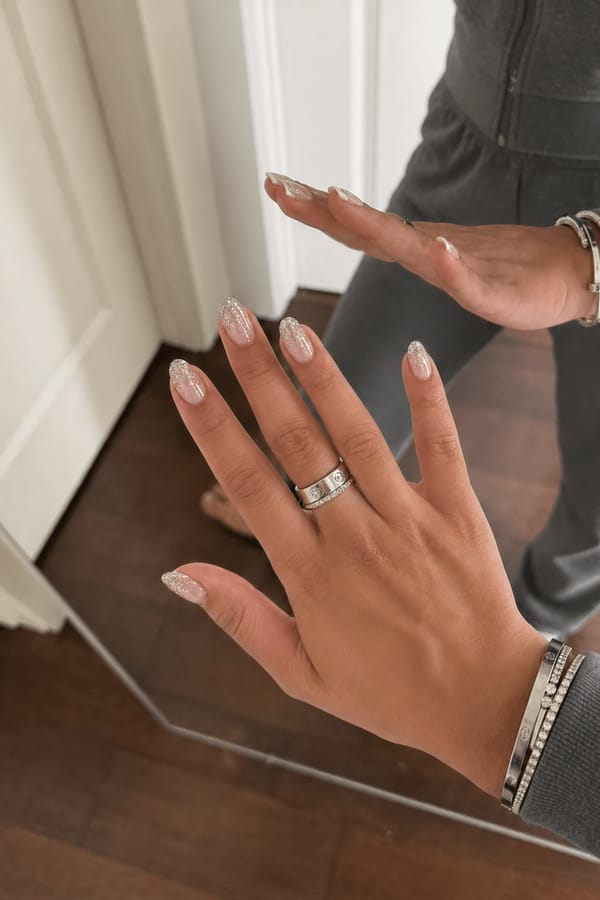 Glossy sheer nude base with a soft ombré fade into fine silver glitter at the tips. 0 accent nails; all nails share the same design. The glitter is densely packed at the tip and gradually diffuses upward into the nude base, creating a smooth, sparkly gradient with no additional details. Nail shape: almond. Nail length: medium. Only change the nails visible in the image to match this design. Keep the rest of the image — the hand, skin, background, lighting, and everything else — exactly the same.