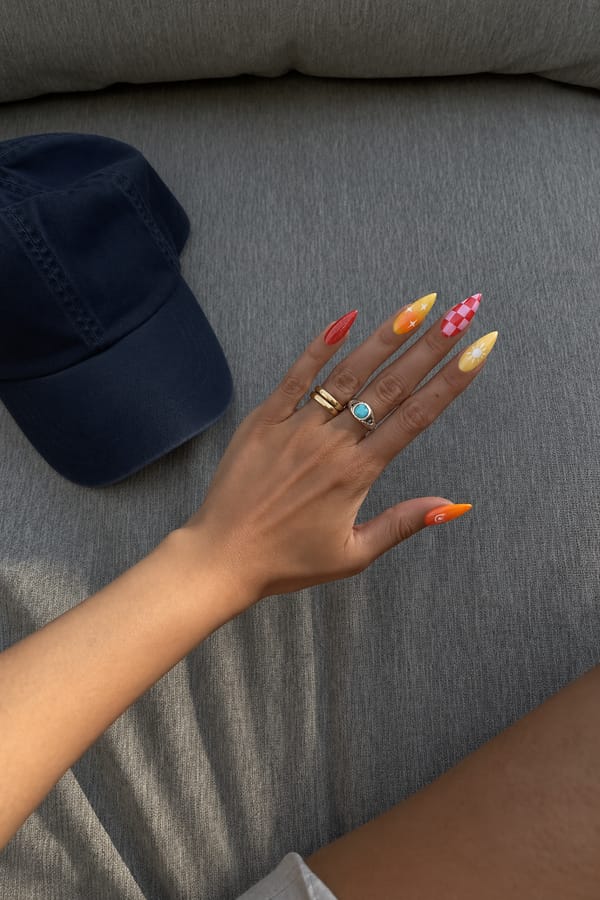 Mixed bright summer set, 5 accent nails: Solid glossy red with fine shimmer. Yellow base with orange aura center and small white star sparkles. Red and light pink checkerboard pattern. Pale yellow base with a white sun motif and radiating curved lines. Solid bright orange base with a small white swirl detail near the tip. Nail shape: stiletto. Nail length: long. Only change the nails visible in the image to match this design. Keep the rest of the image — the hand, skin, background, lighting, and everything else — exactly the same.