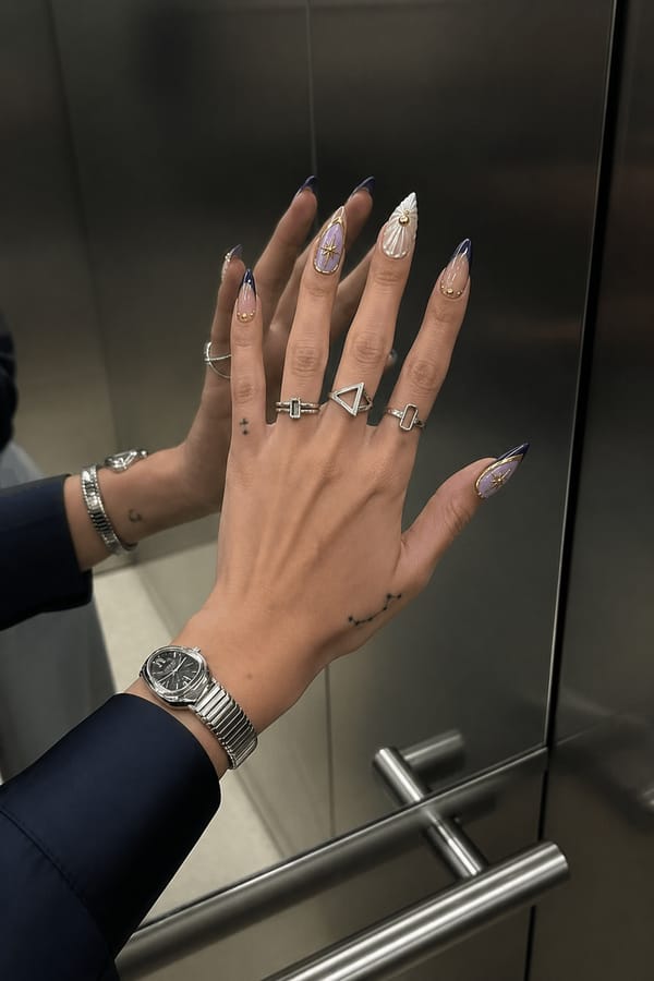 Glossy nude base with deep navy/purple French tips and gold accents. All 5 nails are accent designs. Two nails have soft lavender fill outlined in gold with a central gold starburst. Two nails feature milky white sculpted shell-like patterns with gold studs. One nail has a nude base with a navy tip and small gold studs. Gold details vary per nail. Nail shape: stiletto. Nail length: long. Only change the nails visible in the image to match this design. Keep the rest of the image — the hand, skin, background, lighting, and everything else — exactly the same.