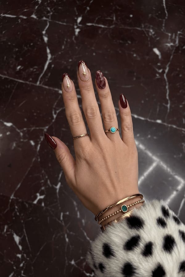 Glossy deep burgundy set with mixed designs. 3 accent nails: one nude base with burgundy French tip edged in gold and a small gold starburst; one nude base with gold outline and starburst center; one translucent burgundy marble effect. Remaining 2 nails are solid glossy burgundy with no design. Nail shape: almond. Nail length: long. Only change the nails visible in the image to match this design. Keep the rest of the image — the hand, skin, background, lighting, and everything else — exactly the same.