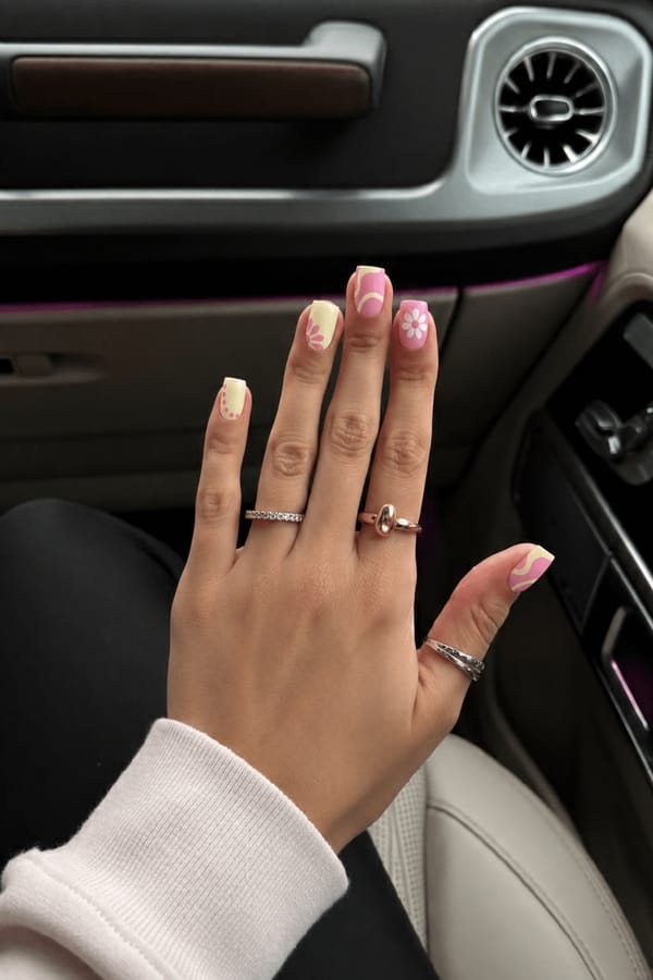 Glossy pastel set, 5 accent nails: Pale yellow base with pink petal burst near one side. Soft pink base with abstract creamy yellow curved shapes. Pink base with simple white flower (5 petals, dotted center). Pale yellow base with pink dotted curve along the edge. Pink base with abstract creamy yellow wave shapes. Nail shape: square. Nail length: medium. Only change the nails visible in the image to match this design. Keep the rest of the image — the hand, skin, background, lighting, and everything else — exactly the same.