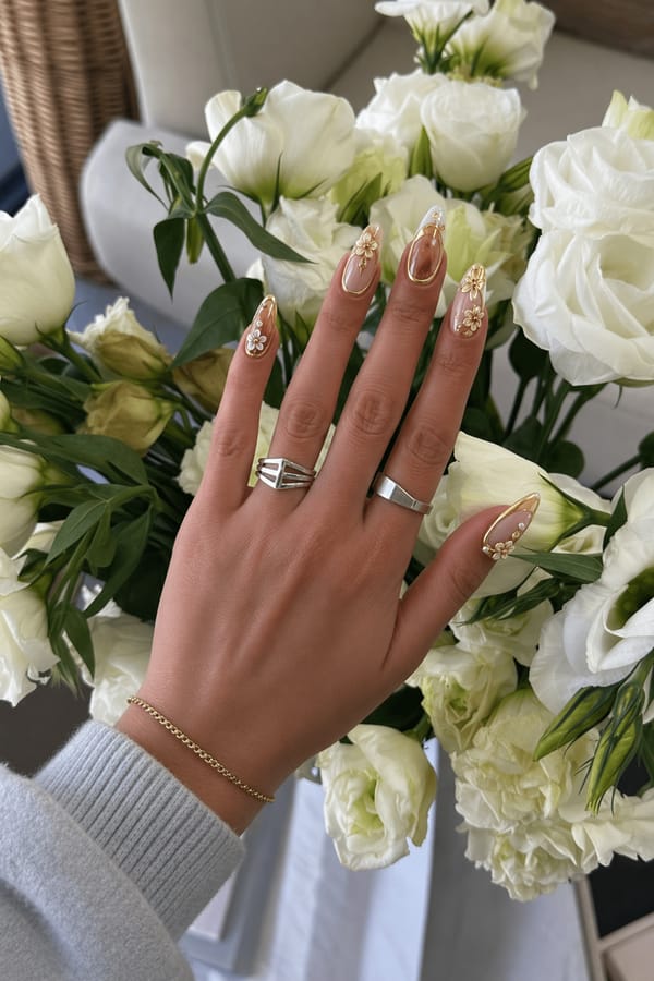 Warm yellow and brown tones with glossy nude accents, gold chrome details, 3D metallic frames, pearl-like elements, and floral embellishments. Luxurious, elegant, high-shine, jewelry-inspired design. Nail shape: almond. Nail length: medium. Only change the nails visible in the image to match this design. Keep the rest of the image — the hand, skin, background, lighting, and everything else — exactly the same.