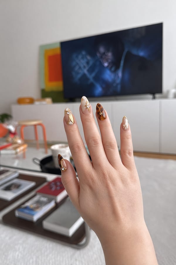 Luxury nail design, long almond nails, mix of glossy tortoiseshell (brown/amber) and milky white + nude bases, minimal negative space. Gold chrome accents, fine metallic lines, small rhinestones, 3D white star embellishments. Elegant, warm-toned, high-gloss finish, editorial style, macro close-up, soft studio lighting. Nail shape: almond. Nail length: long. Only change the nails visible in the image to match this design. Keep the rest of the image — the hand, skin, background, lighting, and everything else — exactly the same.