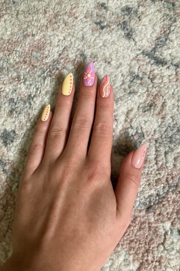 Mixed pastel design with glossy finish. 5 accent nails: Soft yellow base with vertical pink dots along one side. Pink-lilac base with a large 3D-style flower (yellow center). Nude-pink base with abstract hot pink and yellow swirl. Pale yellow base with pink abstract shapes and small white dots. Nude base with minimal pink dot accents near the tip. Nail shape: stiletto. Nail length: medium. Only change the nails visible in the image to match this design. Keep the rest of the image — the hand, skin, background, lighting, and everything else — exactly the same.