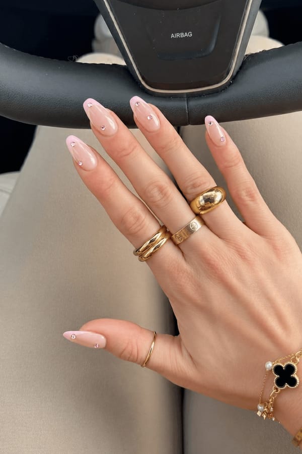 Glossy sheer nude base with soft baby pink French tips. All 5 nails are accent designs: each nail has small light pink rhinestones placed asymmetrically across the surface (center, near tip, or near edges). Variation in placement per nail, but overall minimal scattered crystal design with no additional patterns. Nail shape: almond. Nail length: medium. Only change the nails visible in the image to match this design. Keep the rest of the image — the hand, skin, background, lighting, and everything else — exactly the same.