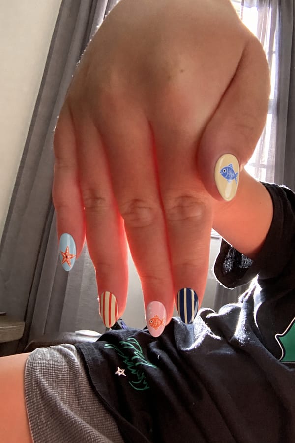 Mixed glossy set, 5 accent nails: Pastel blue base with hand-painted starfish. Nude base with red vertical stripes. Soft pink base with small orange fish illustration. Navy base with beige vertical stripes. Pale yellow base with blue fish illustration. Nail shape: almond. Nail length: short. Only change the nails visible in the image to match this design. Keep the rest of the image — the hand, skin, background, lighting, and everything else — exactly the same.