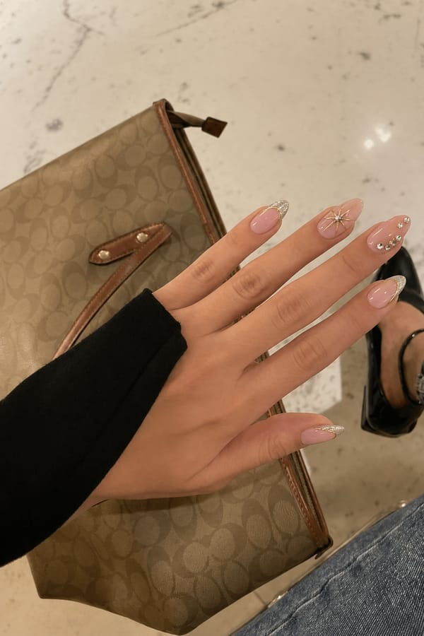 Glossy sheer nude base with mixed designs, 5 accent nails: 1–2) Silver glitter French tips with thin gold outline curves. 3–4) Nude base with central rhinestone and gold starburst lines radiating outward. 5) Nude base with small rhinestone clusters placed asymmetrically near the tip and center. Nail shape: almond. Nail length: medium. Only change the nails visible in the image to match this design. Keep the rest of the image — the hand, skin, background, lighting, and everything else — exactly the same.
