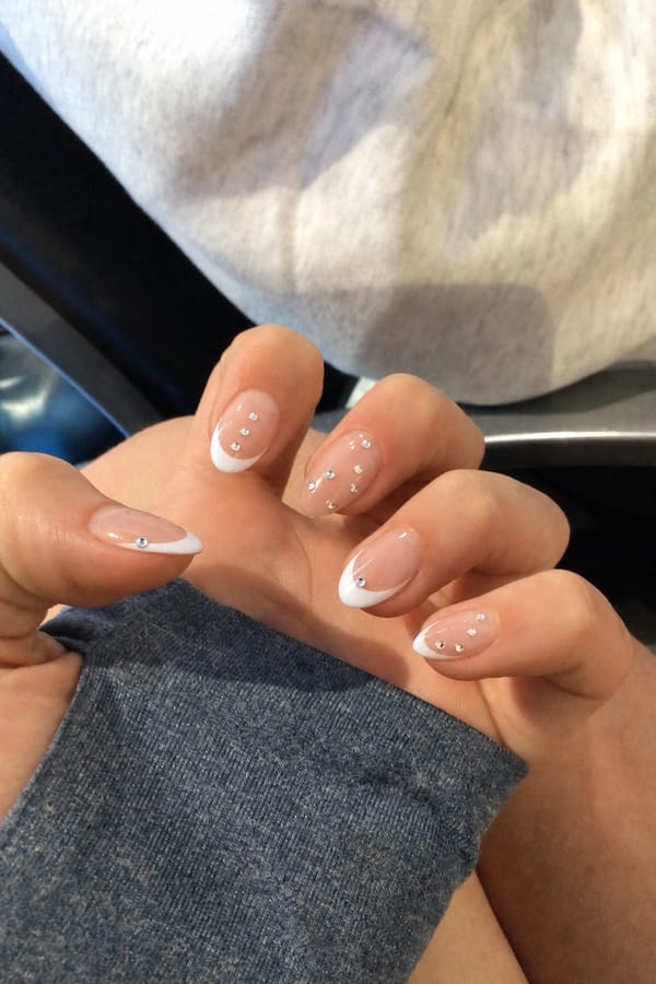 Glossy sheer nude base with crisp white French tips. All 5 nails are accent designs. Each nail features small silver rhinestones placed along the center or near the tip. Variation includes: some nails with classic French tips + scattered rhinestones, some with extended white tips + single rhinestone near the edge, and some nude areas with minimal spaced rhinestones only. Nail shape: almond. Nail length: medium. Only change the nails visible in the image to match this design. Keep the rest of the image — the hand, skin, background, lighting, and everything else — exactly the same.