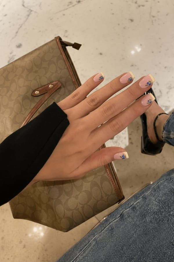Short square nails, nude base with soft pastel yellow French tips, tiny blueberry nail art with green stems. Cute, fresh, minimal aesthetic, glossy finish Nail shape: square. Nail length: medium. Only change the nails visible in the image to match this design. Keep the rest of the image — the hand, skin, background, lighting, and everything else — exactly the same.