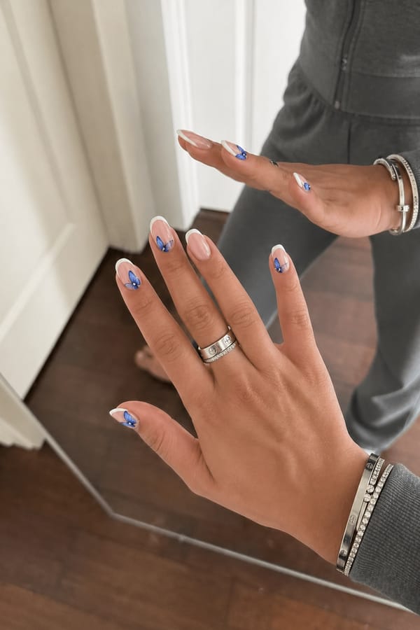 Glossy nude base with crisp white French tips on all nails. 3 accent nails: each features a blue butterfly design with two gradient blue wings and a small silver bead center, placed near the tip or slightly off-center. Remaining 2 nails have clean white French tips only with no additional design. Nail shape: almond. Nail length: medium. Only change the nails visible in the image to match this design. Keep the rest of the image — the hand, skin, background, lighting, and everything else — exactly the same.