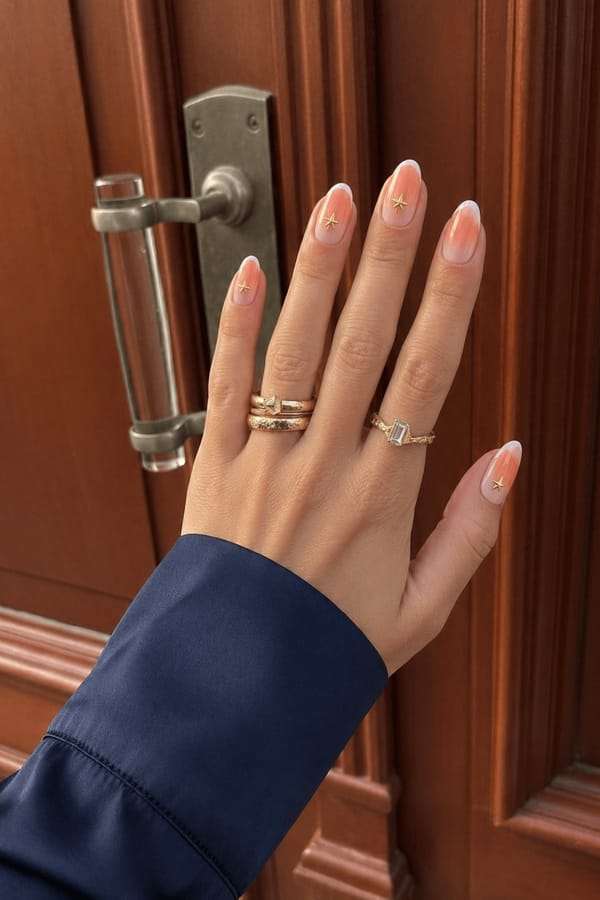 Sheer nude glossy base with a soft orange-to-white radial gradient (aura effect) concentrated toward the tips. A mix of styles across the set: some nails feature a smooth gradient only, while others include blended white French tips layered over the gradient. Accent details on 4 nails: 3 nails have small gold star decals placed slightly above the tip area, centered or slightly offset. 1 nail has a gold star decal placed lower toward the mid-area of the nail. Nail shape: almond. Nail length: medium. Only change the nails visible in the image to match this design. Keep the rest of the image — the hand, skin, background, lighting, and everything else — exactly the same.