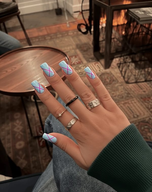 Nails featuring a soft pastel palette with a base color of light blue across most nails, complemented by delicate geometric patterns in pink and lilac. The index and ring fingers showcase a minimalist design with intersecting triangles and thin lines creating a modern, abstract look, while the other nails remain a smooth, solid light blue. The geometric elements are kept subtle and sparse, with no more than four shapes per accented nail, ensuring an elegant and feminine finish. A glossy top coat enhances the overall appearance, adding a fresh and airy vibe to these medium-length square nails.