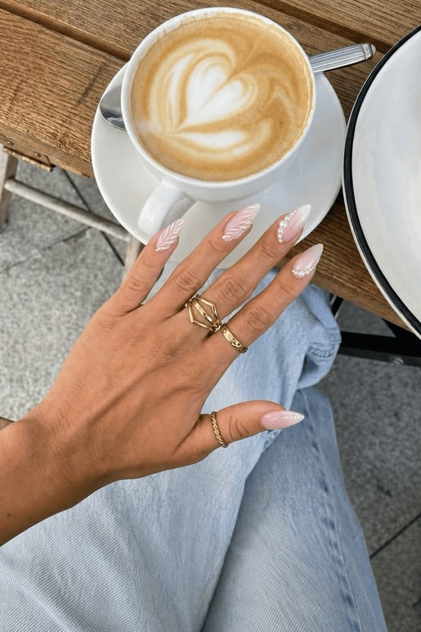 Glossy sheer nude base with milky white gradients. 5 accent nails: 1–2) White feather-like 3D sculpted strokes flowing diagonally. 3) Soft white gradient with a curved line of small pearls along one side. 4) Feather-like 3D strokes plus a single pearl cluster near the center. 5) White gradient with minimal pearl accents near the edge. Nail shape: stiletto. Nail length: medium. Only change the nails visible in the image to match this design. Keep the rest of the image — the hand, skin, background, lighting, and everything else — exactly the same.