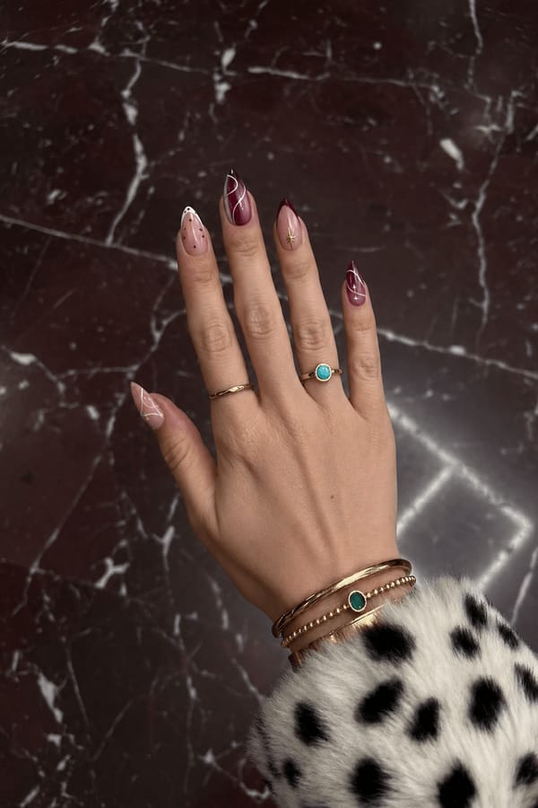 Glossy sheer pink base with burgundy tones. 5 accent nails: Nude base with white tip and small burgundy dots. Burgundy aura/gradient with fine white curved lines. Nude base with burgundy French tip and small gold star detail. Burgundy gradient with white swirl lines and subtle star accent. Nude base with thin abstract white lines across the surface. Nail shape: stiletto. Nail length: long. Only change the nails visible in the image to match this design. Keep the rest of the image — the hand, skin, background, lighting, and everything else — exactly the same.