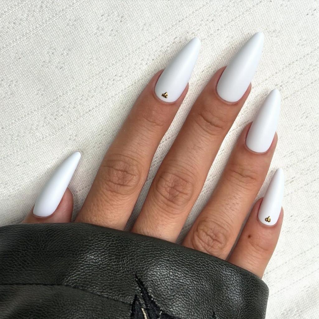 Long stiletto nails in matte white, each featuring a small gold stud near the cuticle.