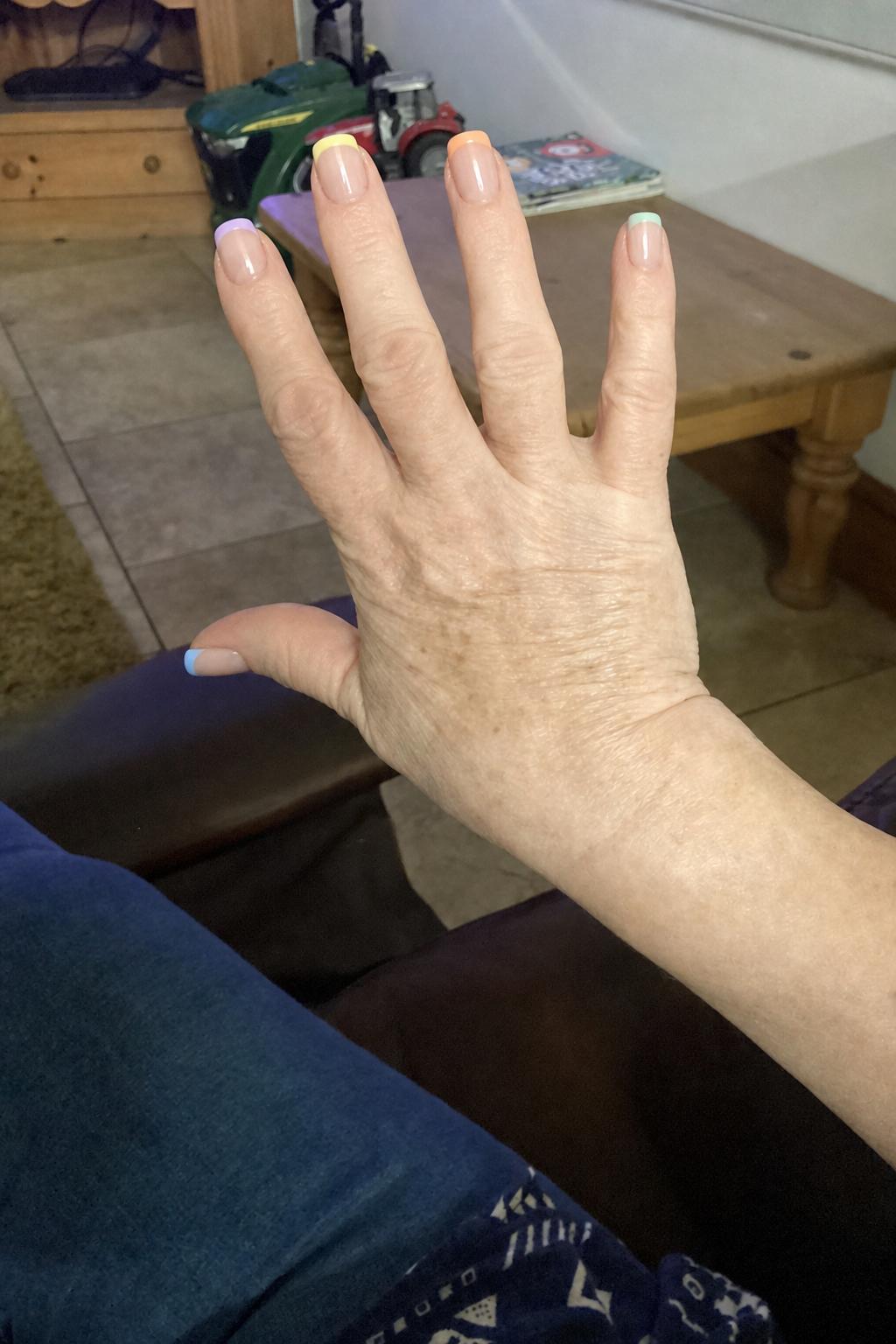 Glossy sheer nude base with pastel French tips. 5 accent nails: each nail features a different soft pastel tip color (lavender, yellow, peach, pink, mint, baby blue tones). Tips are clean and minimal with no outlines or decorations. All nails share the same style, only color varies per nail. Nail shape: square. Nail length: medium. Only change the nails visible in the image to match this design. Keep the rest of the image — the hand, skin, background, lighting, and everything else — exactly the same.