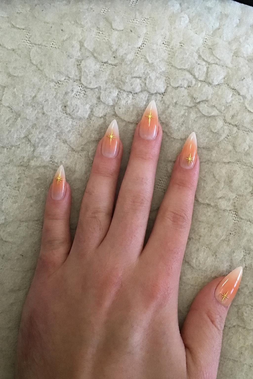 Sheer nude glossy base with soft orange-to-white radial gradient (aura) focused toward the tips. Some nails have blended white French tips over the gradient. 4 accent nails: 3 with small gold star decals near the tip, 1 with a gold star placed slightly lower. Remaining nails are gradient-only or gradient + soft French blend. Nail shape: stiletto. Nail length: long. Only change the nails visible in the image to match this design. Keep the rest of the image — the hand, skin, background, lighting, and everything else — exactly the same.