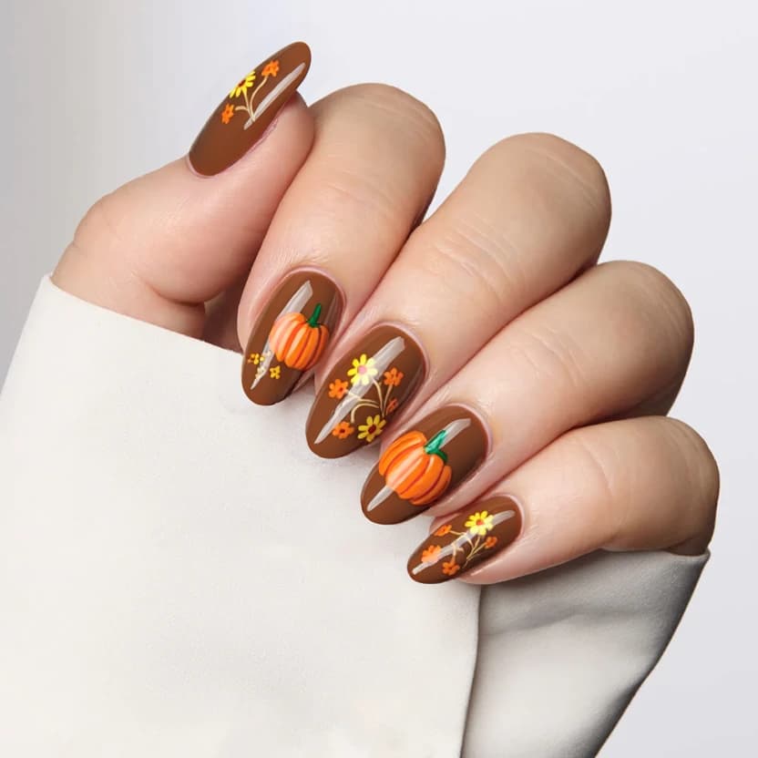 Nails featuring a warm, creamy brown base, designed to evoke a cozy autumn feel. The tips of the almond-shaped nails are adorned with delicate, hand-painted orange pumpkins, artistically placed to add a touch of seasonal charm without overwhelming the design. Accentuating the pumpkins, small yellow sunflowers and dainty orange daisies are strategically scattered on two of the nails, creating a harmonious, floral composition. The remaining nails maintain the soft brown background, ensuring a balanced and elegant look. A glossy finish enhances the overall appearance, bringing to life the intricate details while keeping the design sophisticated and feminine.