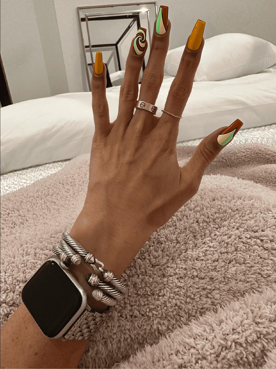 Nails featuring a soft, creamy base color that complements the retro aesthetic, with three accent nails showcasing a playful design. On the middle finger of one hand, a swirling pattern in burnt orange and avocado green flows elegantly from the base to the tip, creating a dynamic movement. The ring finger presents a geometric pattern with bold, angular shapes in mustard yellow and burnt orange, adding a touch of retro flair. The remaining nails are kept in a solid creamy base color, ensuring a harmonious balance between the intricate designs and the simplicity of the background. A glossy finish enhances the vibrant colors, perfectly embodying the playful spirit of the 70s while fitting beautifully on medium-length almond-shaped nails.