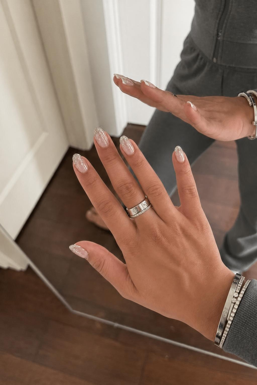 Glossy sheer nude base with a soft ombré fade into fine silver glitter at the tips. 0 accent nails; all nails share the same design. The glitter is densely packed at the tip and gradually diffuses upward into the nude base, creating a smooth, sparkly gradient with no additional details. Nail shape: almond. Nail length: medium. Only change the nails visible in the image to match this design. Keep the rest of the image — the hand, skin, background, lighting, and everything else — exactly the same.