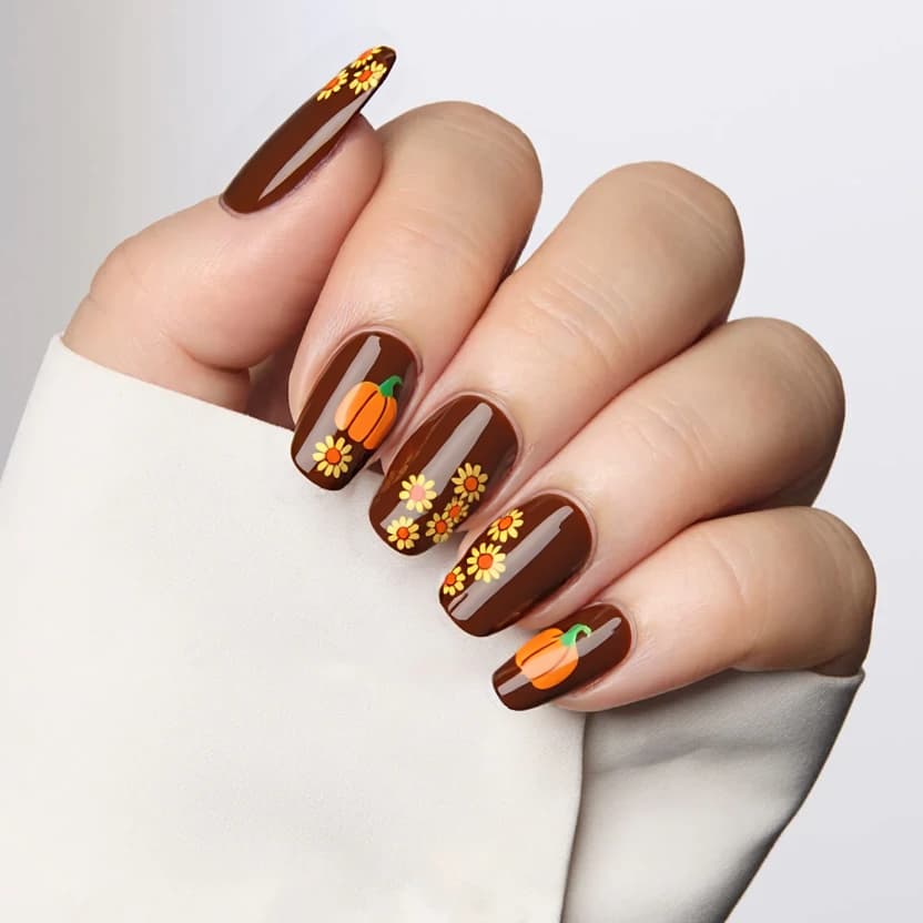 Nails painted in a rich, warm brown to create an elegant base, complementing the almond shape and medium size. The tips of the nails are adorned with charming, minimalistic orange pumpkins, delicately positioned to add a playful touch. Accenting this autumn-inspired design, small yellow sunflowers and vivid orange daisies are tastefully scattered across two of the nails, enhancing the overall feminine feel while maintaining a simple aesthetic. The remaining nails are kept plain brown, offering a harmonious balance to the design. A glossy finish brings a subtle shine, giving the nails a polished, seasonal look.