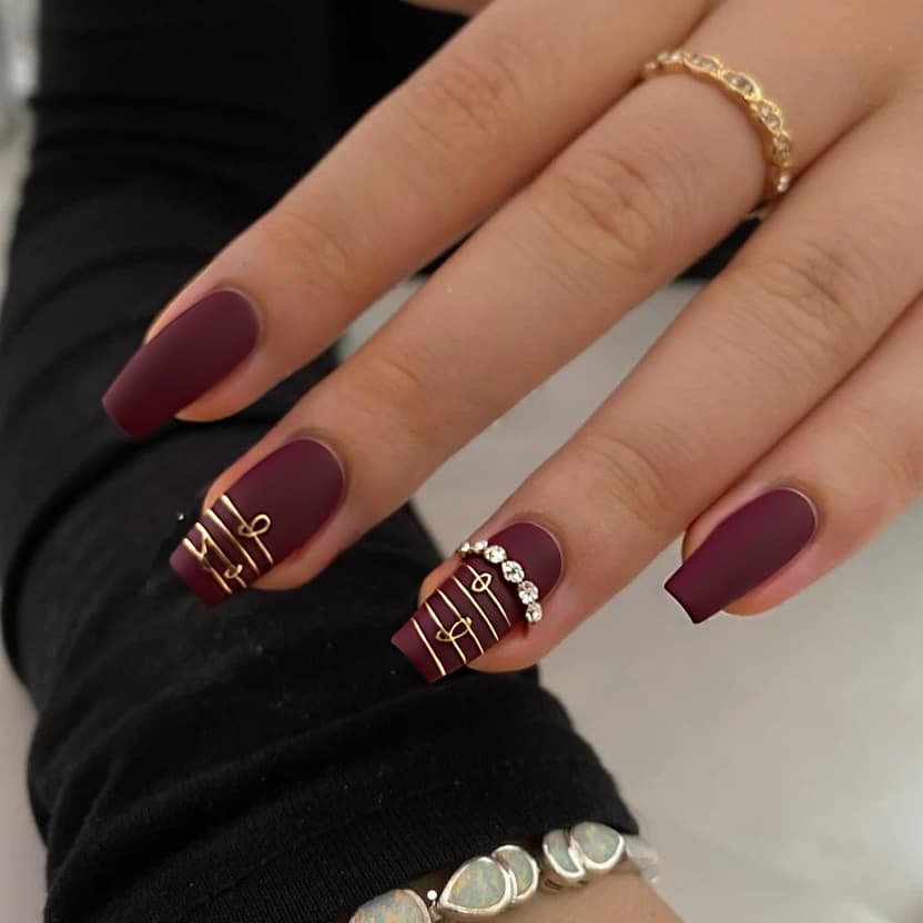 Short stiletto nails coated in a deep, dark red base that exudes elegance. On the ring fingers, a delicate design of minimalist music notes, hand-painted in subtle metallic gold, adds a unique touch. These notes are gently placed to flow diagonally across the nail, while the other fingers feature a smooth, solid dark red finish. A few small crystal accents are carefully placed near the base of the music notes for a touch of sparkle, creating a harmonious blend of sophistication and playful charm. The overall effect is chic and refined, perfectly suited for the sleek lines and pointed tips of short stiletto nails.
