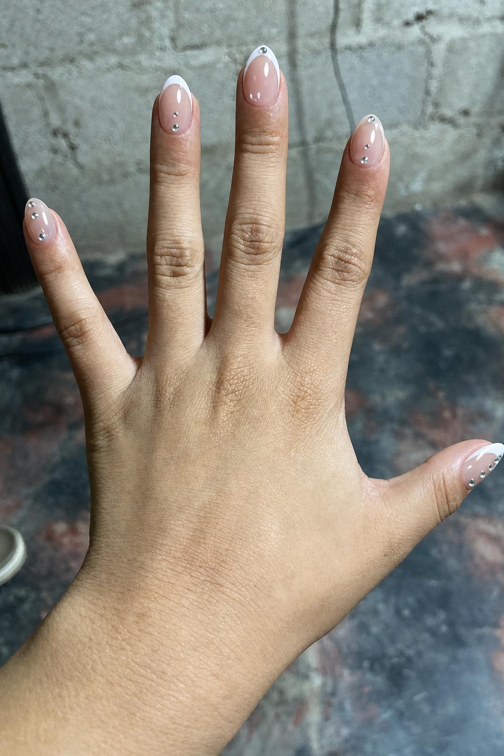 Glossy sheer nude base with crisp white French tips. All 5 nails are accent designs. Each nail features small silver rhinestones placed along the center or near the tip. Variation includes: some nails with classic French tips + scattered rhinestones, some with extended white tips + single rhinestone near the edge, and some nude areas with minimal spaced rhinestones only. Nail shape: almond. Nail length: medium. Only change the nails visible in the image to match this design. Keep the rest of the image — the hand, skin, background, lighting, and everything else — exactly the same.