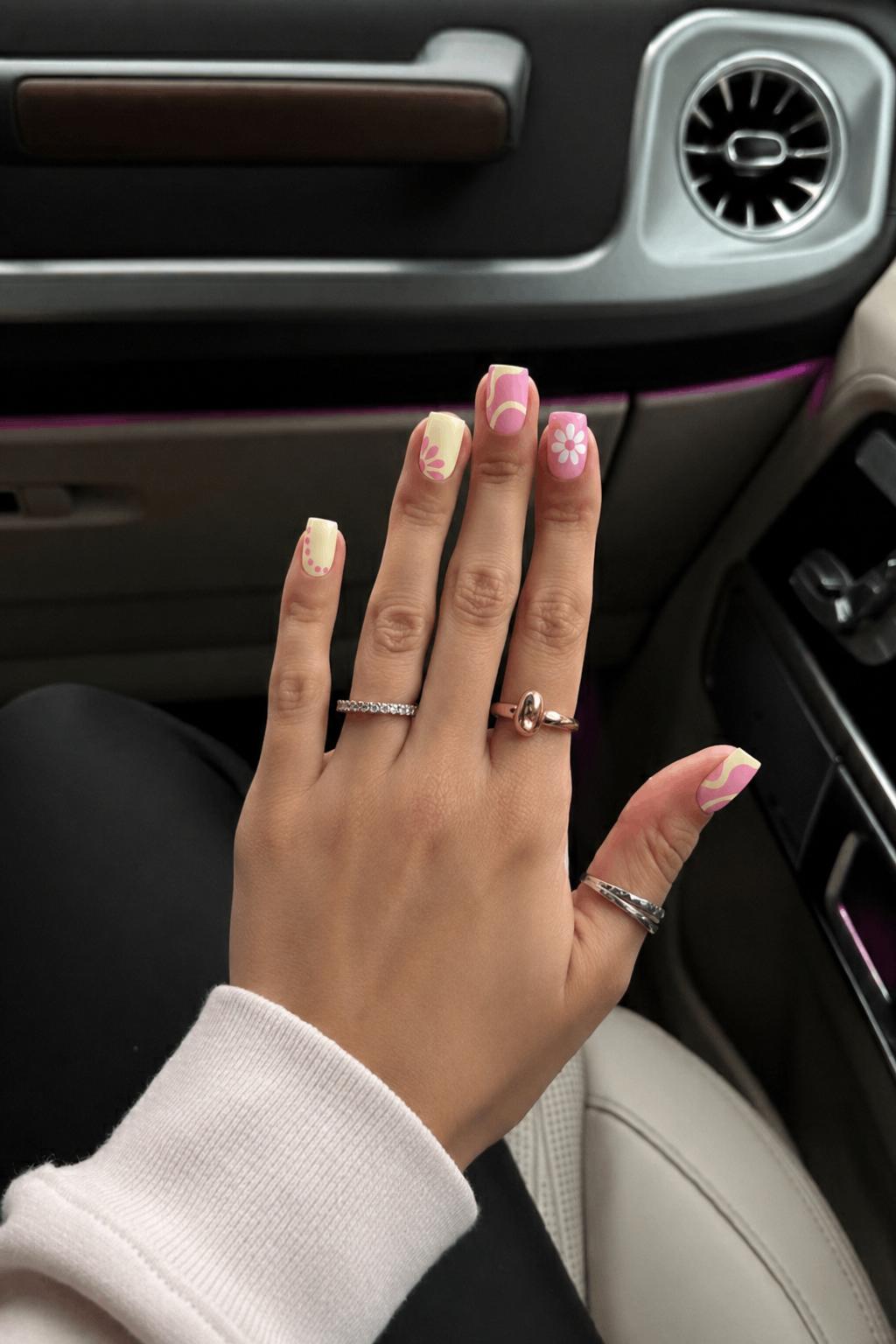 Glossy pastel set, 5 accent nails: Pale yellow base with pink petal burst near one side. Soft pink base with abstract creamy yellow curved shapes. Pink base with simple white flower (5 petals, dotted center). Pale yellow base with pink dotted curve along the edge. Pink base with abstract creamy yellow wave shapes. Nail shape: square. Nail length: medium. Only change the nails visible in the image to match this design. Keep the rest of the image — the hand, skin, background, lighting, and everything else — exactly the same.