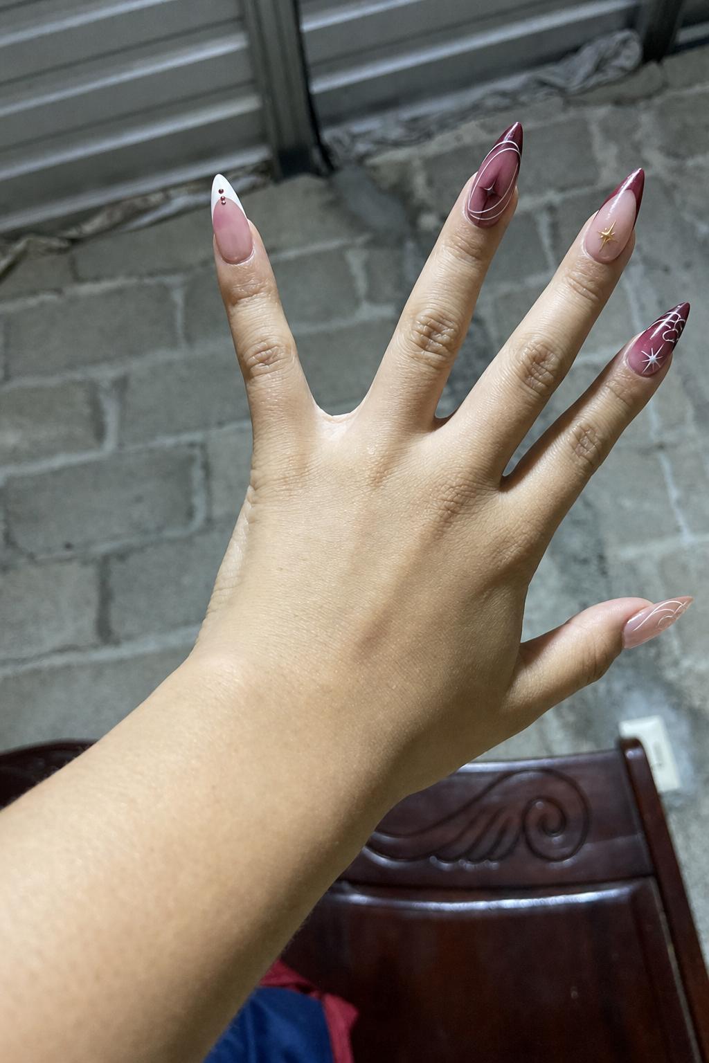Glossy sheer pink base with burgundy tones. 5 accent nails: Nude base with white tip and small burgundy dots. Burgundy aura/gradient with fine white curved lines. Nude base with burgundy French tip and small gold star detail. Burgundy gradient with white swirl lines and subtle star accent. Nude base with thin abstract white lines across the surface. Nail shape: stiletto. Nail length: long. Only change the nails visible in the image to match this design. Keep the rest of the image — the hand, skin, background, lighting, and everything else — exactly the same.