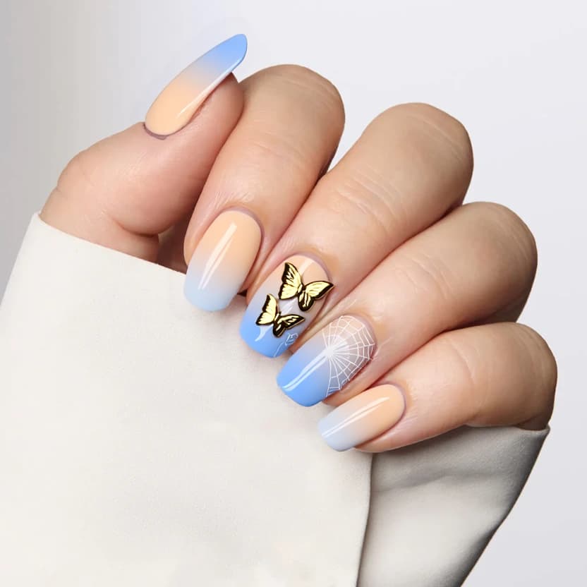 Nails featuring a serene baby boomer gradient, transitioning softly from a warm beige base to a delicate pastel blue at the tips. The design enhances the elegant almond shape of the nails. On the ring finger, a lovely arrangement of golden butterfly stickers is artfully placed, beginning near the base of the nail and gracefully trailing towards the tip, adding a touch of whimsy. On the middle finger, a subtle white spider gel effect is layered above the baby boomer, creating an intricate and airy pattern that complements the overall design without overwhelming it. The finish is glossy, enhancing the gentle hues and details, making for a chic and feminine look perfect for medium almond nails.