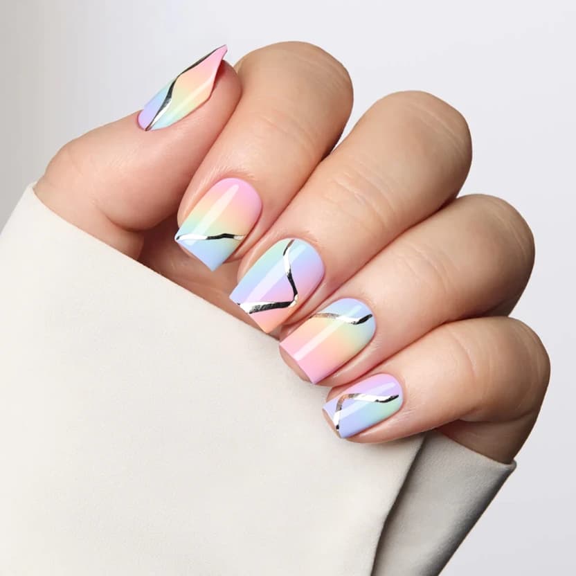 Nails in a soft, pastel gradient inspired by a colorful sunset, transitioning seamlessly from a light peach to a delicate lavender and a hint of soft blue. The base color is consistent across all nails, creating a serene backdrop. On the ring fingers, minimalistic metallic silver accents create elegant, abstract lines that intertwine gently with the gradient, providing a subtle contrast without overwhelming the design. The overall finish is glossy, enhancing the warm tones while ensuring the design remains modern and sophisticated, perfectly suited for the short square shape.