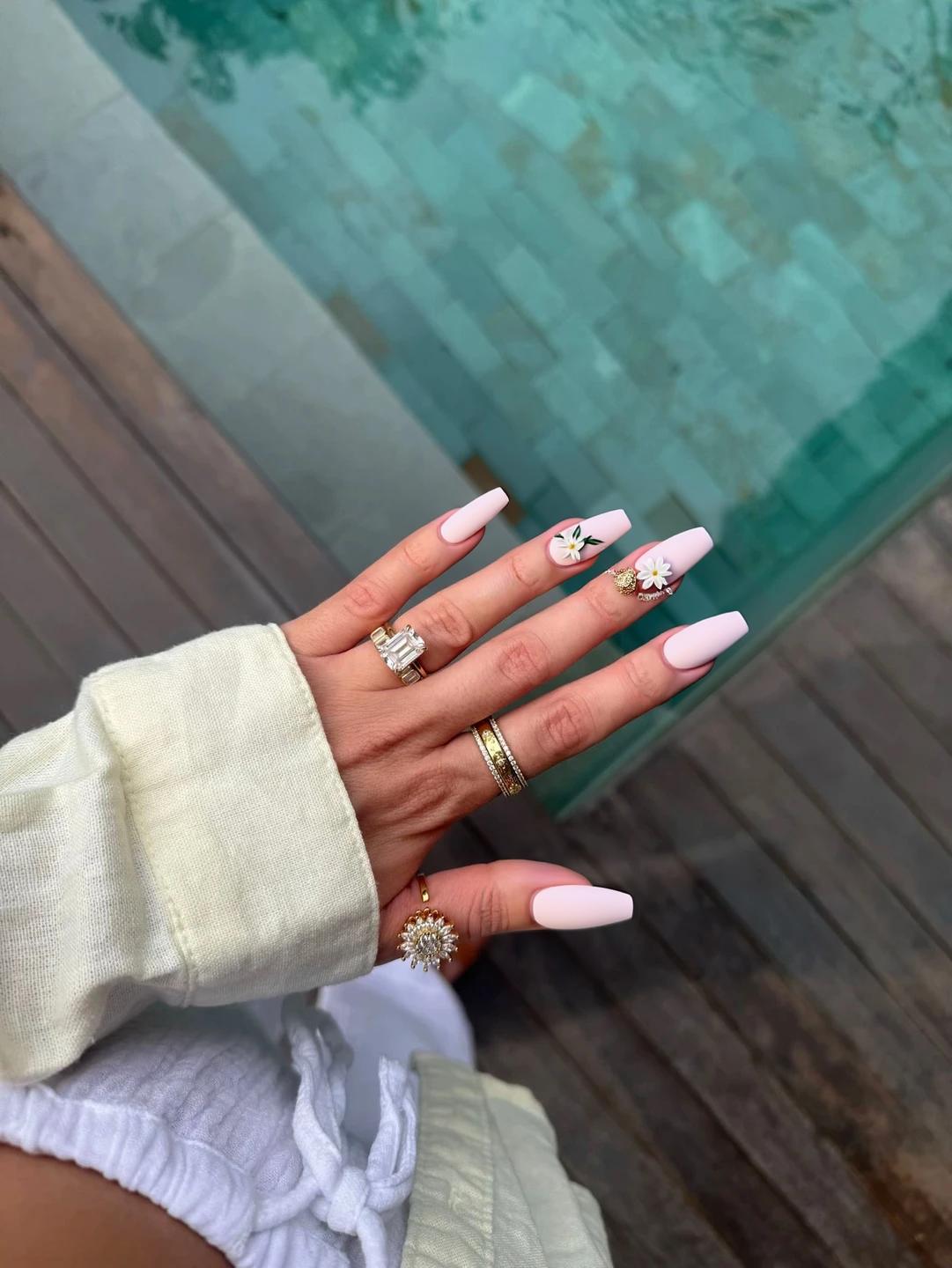 Nails adorned with a soft blush pink base, creating a romantic and gentle aesthetic ideal for a summer wedding. The ring fingers feature delicate white floral accents, with minimal hand-painted blossoms and tiny green leaves, maintaining an elegant and understated design. The other nails are kept plain in the same soft pink hue to enhance the overall harmony of the look. A glossy finish adds a touch of sophistication, perfectly complementing the oval shape and medium length of the nails, ensuring a graceful appearance for the occasion.