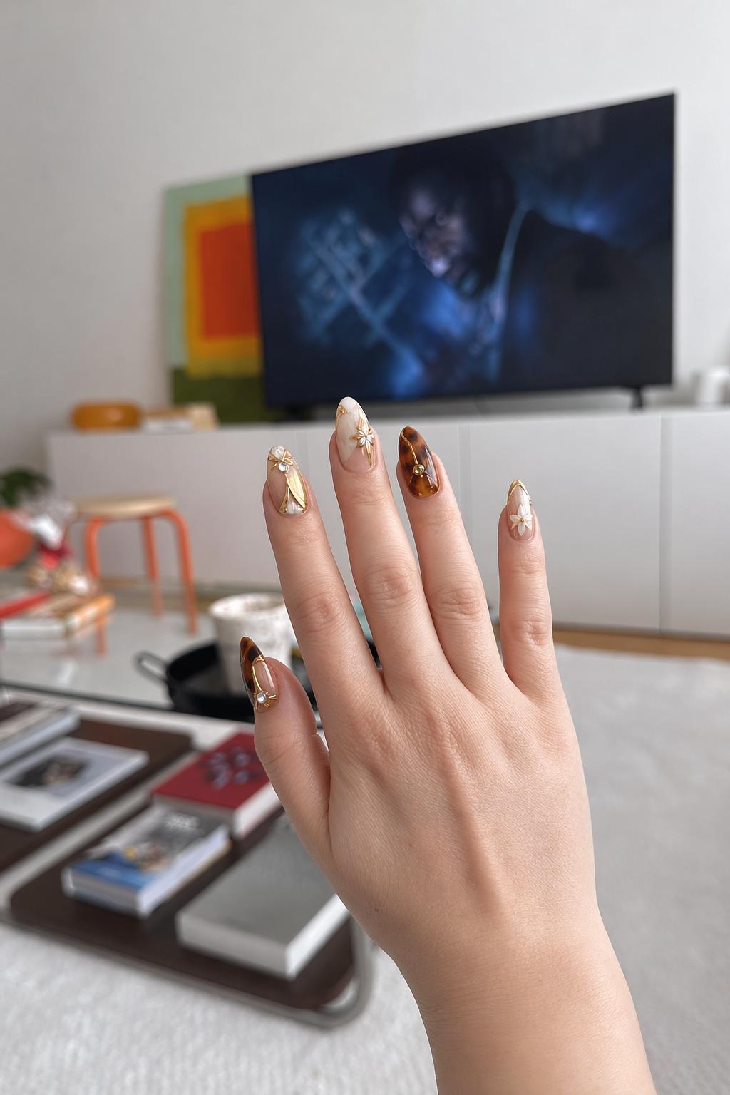 Luxury nail design, long almond nails, mix of glossy tortoiseshell (brown/amber) and milky white + nude bases, minimal negative space. Gold chrome accents, fine metallic lines, small rhinestones, 3D white star embellishments. Elegant, warm-toned, high-gloss finish, editorial style, macro close-up, soft studio lighting. Nail shape: almond. Nail length: long. Only change the nails visible in the image to match this design. Keep the rest of the image — the hand, skin, background, lighting, and everything else — exactly the same.
