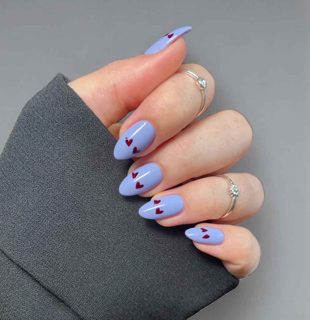 Nails in a soft pastel base color create an elegant backdrop, enhancing the almond shape's graceful curves. Short almond nails feature a delicate dark blue French tip that adds a touch of sophistication to the manicure. The rest of the nail is adorned with small, minimalistic dark red hearts, placed thoughtfully to maintain a balanced and refined appearance. The overall finish is glossy, which accentuates the rich colors while providing a sleek look that is both feminine and chic.