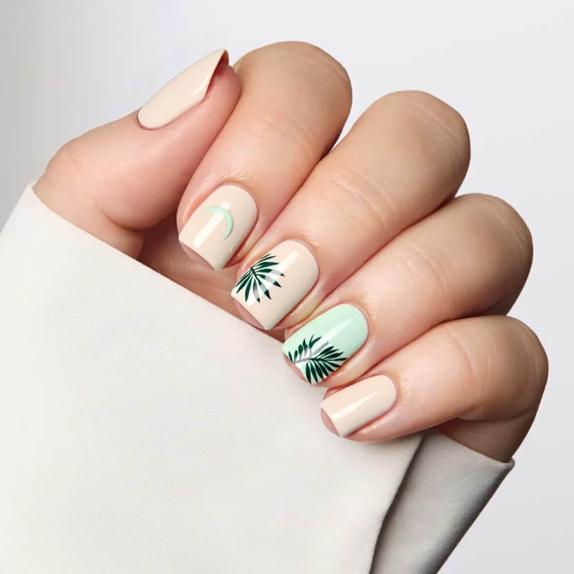 Short square nails featuring a tranquil beachy vibe, painted in a soft, sandy beige as the base color to evoke the essence of a beach shore. On the accent nails—the ring fingers—delicate, hand-painted tropical leaves in a subtle mint green gently arch across, creating an elegant yet playful contrast against the neutral background. The remaining nails are left in a plain sandy beige, maintaining a minimalistic aesthetic. A glossy finish adds a fresh, sun-kissed feel to the overall design, perfectly suited for the simple yet chic nature of short square nails.