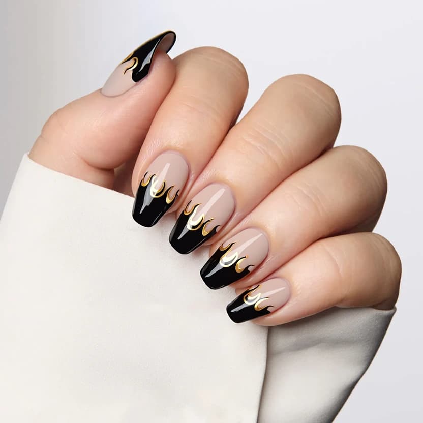 Nails feature a sheer nude base, providing an elegant and understated foundation. The almond-shaped nails are adorned with sharp black French tips that create a striking contrast, emphasizing the gentle curve of the nail's edge. On the ring fingers, delicate accents of small gold foil flames gracefully rise, adding a touch of flamboyance without overwhelming the design. The overall finish is glossy, enhancing the sophisticated look while maintaining a feminine charm that perfectly complements the medium length of the nails.