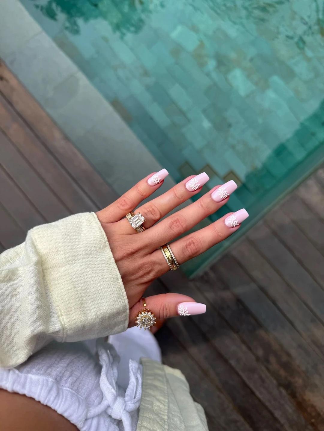 Nails featuring a soft pink base, creating a romantic and delicate look perfect for a summer wedding. On the ring fingers, subtle white floral accents are hand-painted, consisting of tiny blossoms and delicate leaves, arranged in a minimalist style with no more than four elements per nail. The remaining nails maintain the soft pink hue, providing a harmonious and elegant backdrop. A glossy finish enhances the overall femininity of the design, making it sophisticated yet understated, ideal for a wedding setting.