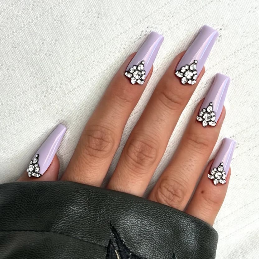 Long stiletto nails will be adorned in a soft, pastel lilac base color, creating an elegant and feminine atmosphere. Each nail features a minimalistic arrangement of sparkling crystal rhinestones, with no more than three strass placed delicately along the cuticle line, providing a hint of glamour without overwhelming the design. The design maintains a glossy finish to enhance the shine of the rhinestones and the overall sophisticated look, perfectly suited for the dramatic and elongated shape of stiletto nails.