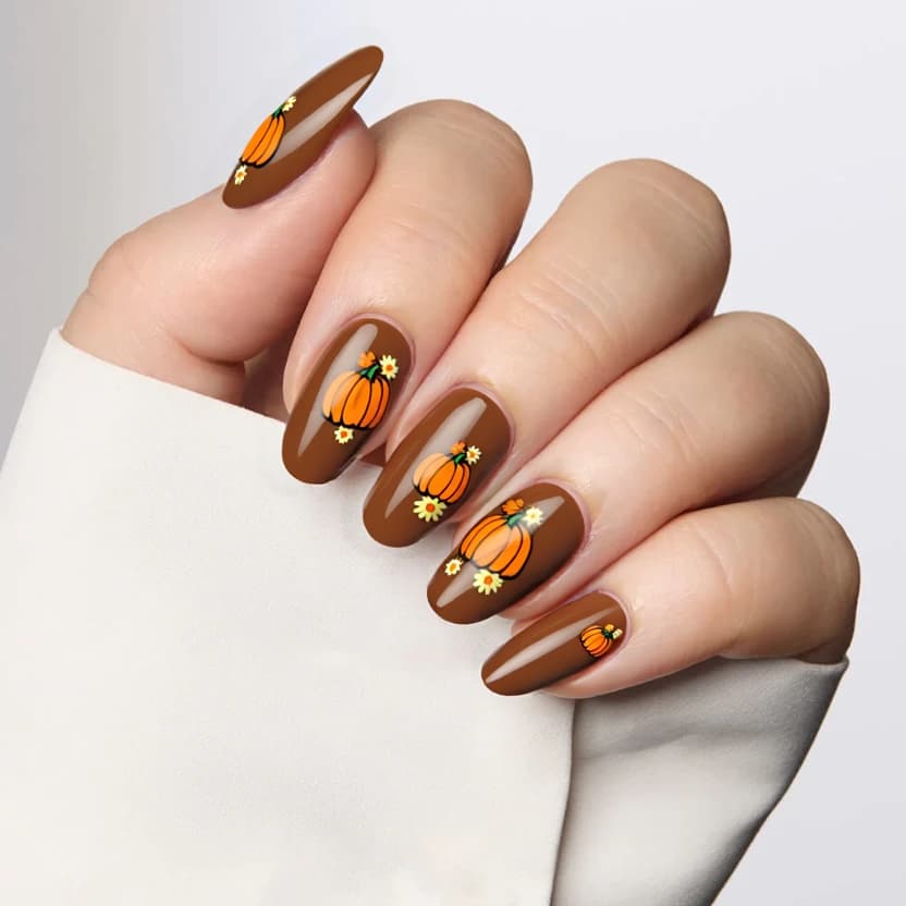 Nails featuring a smooth, warm brown base color that creates an inviting and earthy backdrop. The tips of each nail are adorned with minimalistic depictions of small orange pumpkins, maintaining a delicate and playful feel that suits the almond shape beautifully. On the ring fingers, small yellow sunflowers and orange daisies are tastefully arranged, adding a cheerful accent without overwhelming the design. The artwork on the nails is kept minimal, ensuring that it is elegant and feminine, while the other nails remain a flawless brown, providing a cohesive look. A glossy finish enhances the overall aesthetic, giving the nails a polished and vibrant appearance.